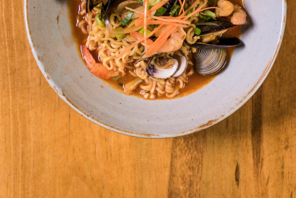 Top view of a tasty seafood ramen dish with shrimp, mussels, and clams in a ceramic bowl on wooden surface.