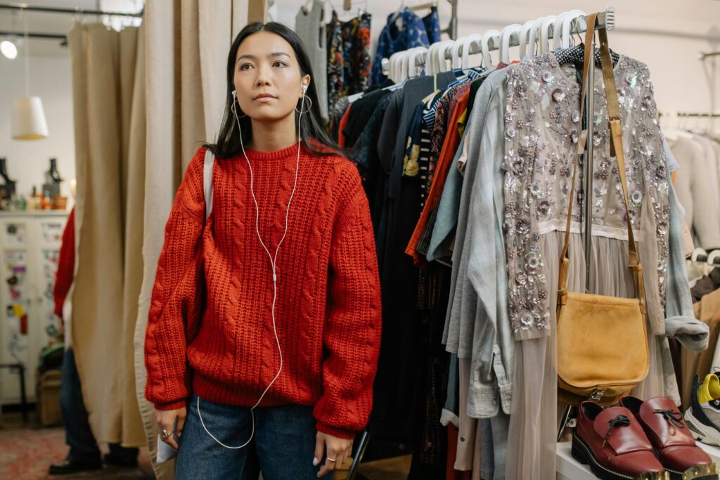 Asian woman in red sweater shopping, listening to music in a clothing store. Stylish and trendy fashion choices.